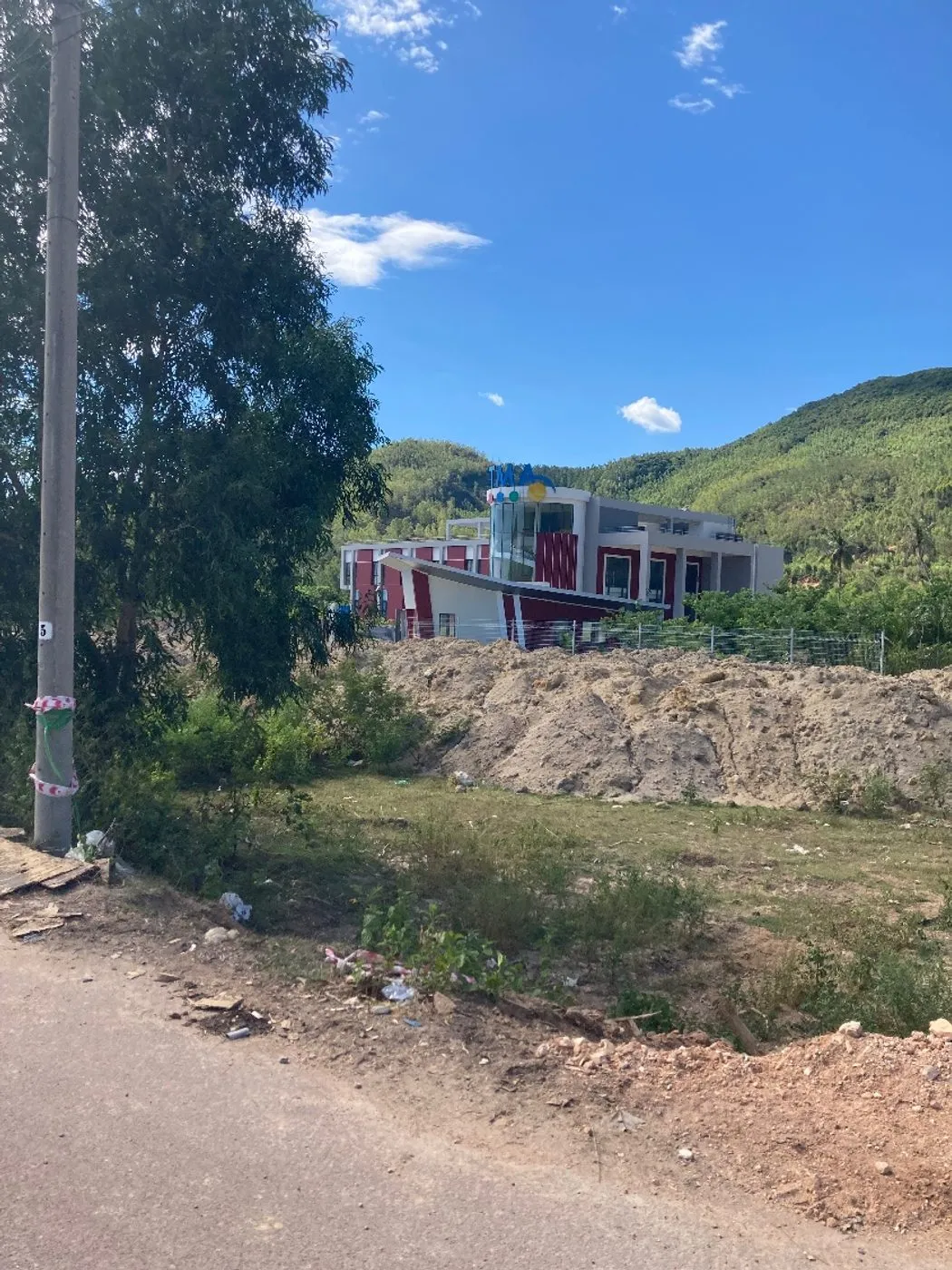 A modern red-and-white building with a glass tower, fenced off behind mounds of dumped earth and a rutted road with litter in the verge, green hills behind.