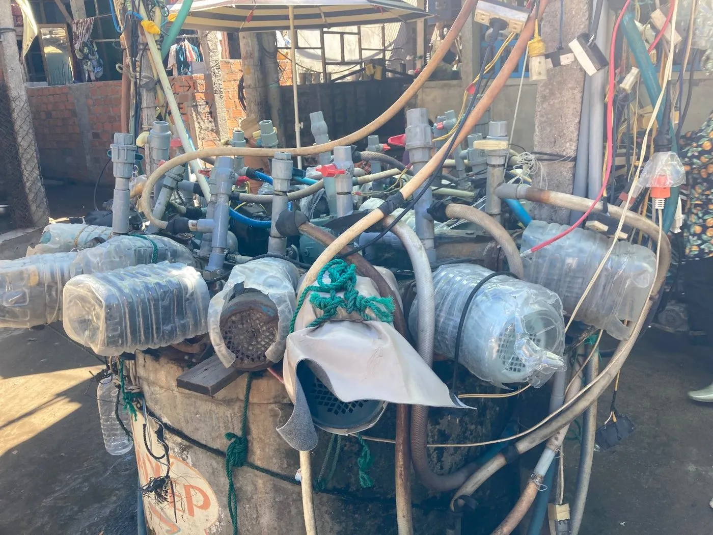 A round concrete well crowded with a dozen electric pumps, each with its own grey pipe, hose and wiring; many motors are wrapped in cut-off plastic water bottles, one covered with a scrap of tarpaulin tied on with green rope.
